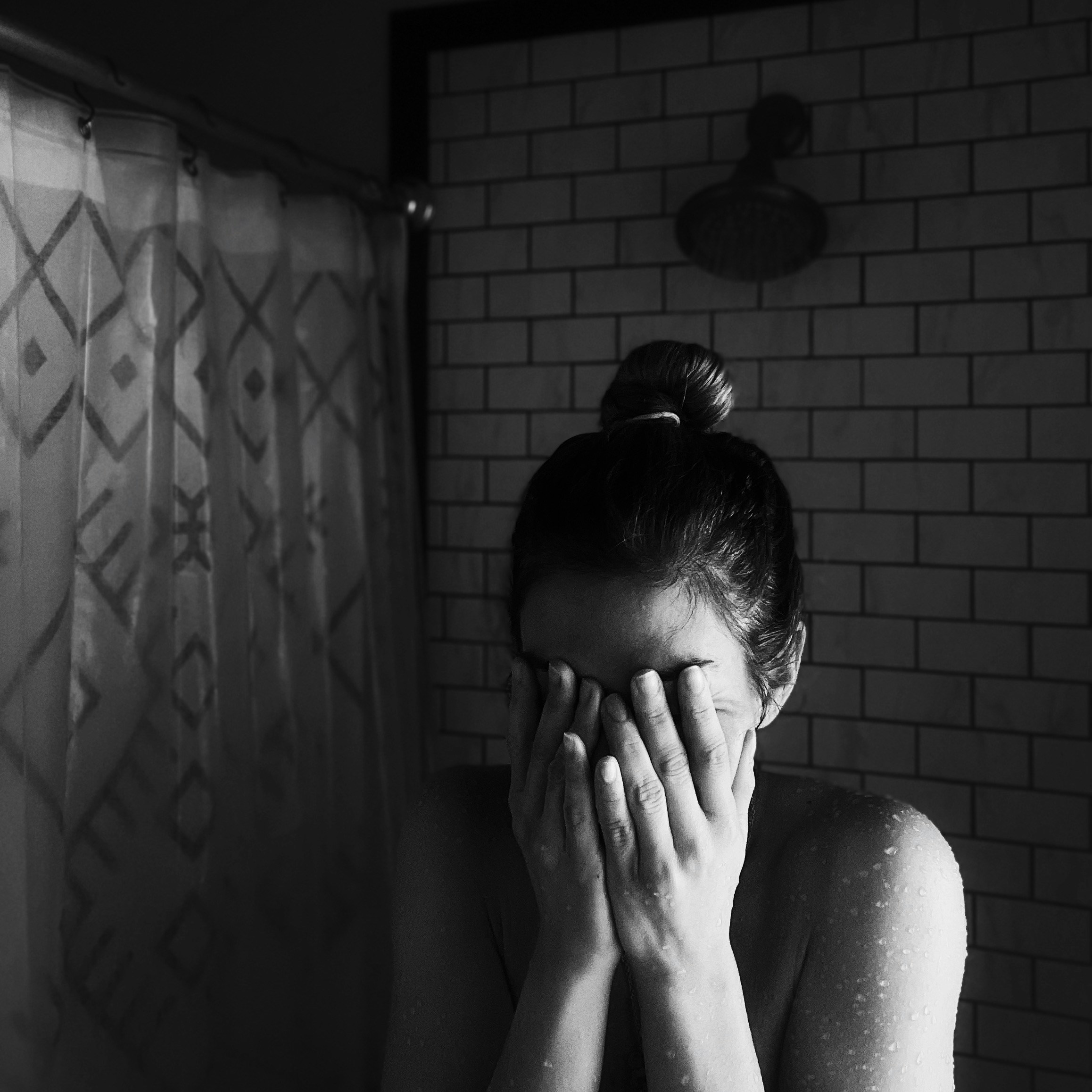 A woman standing under a shower, covering her face with both hands, expressing emotional distress or anxiety, in a dark and moody bathroom setting.