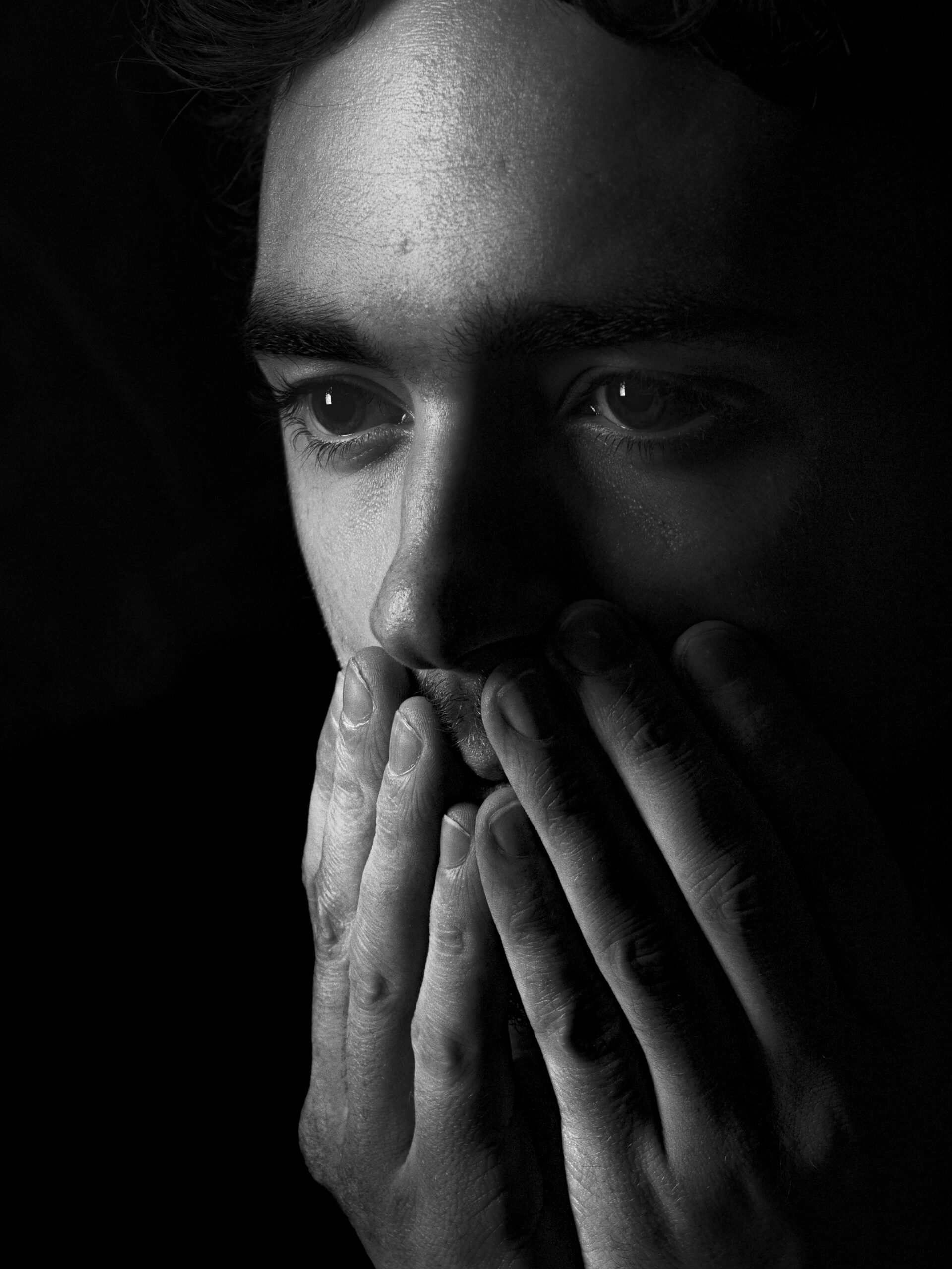 A close-up black and white portrait of a man covering his mouth with both hands, expressing deep anxiety or emotional distress.
